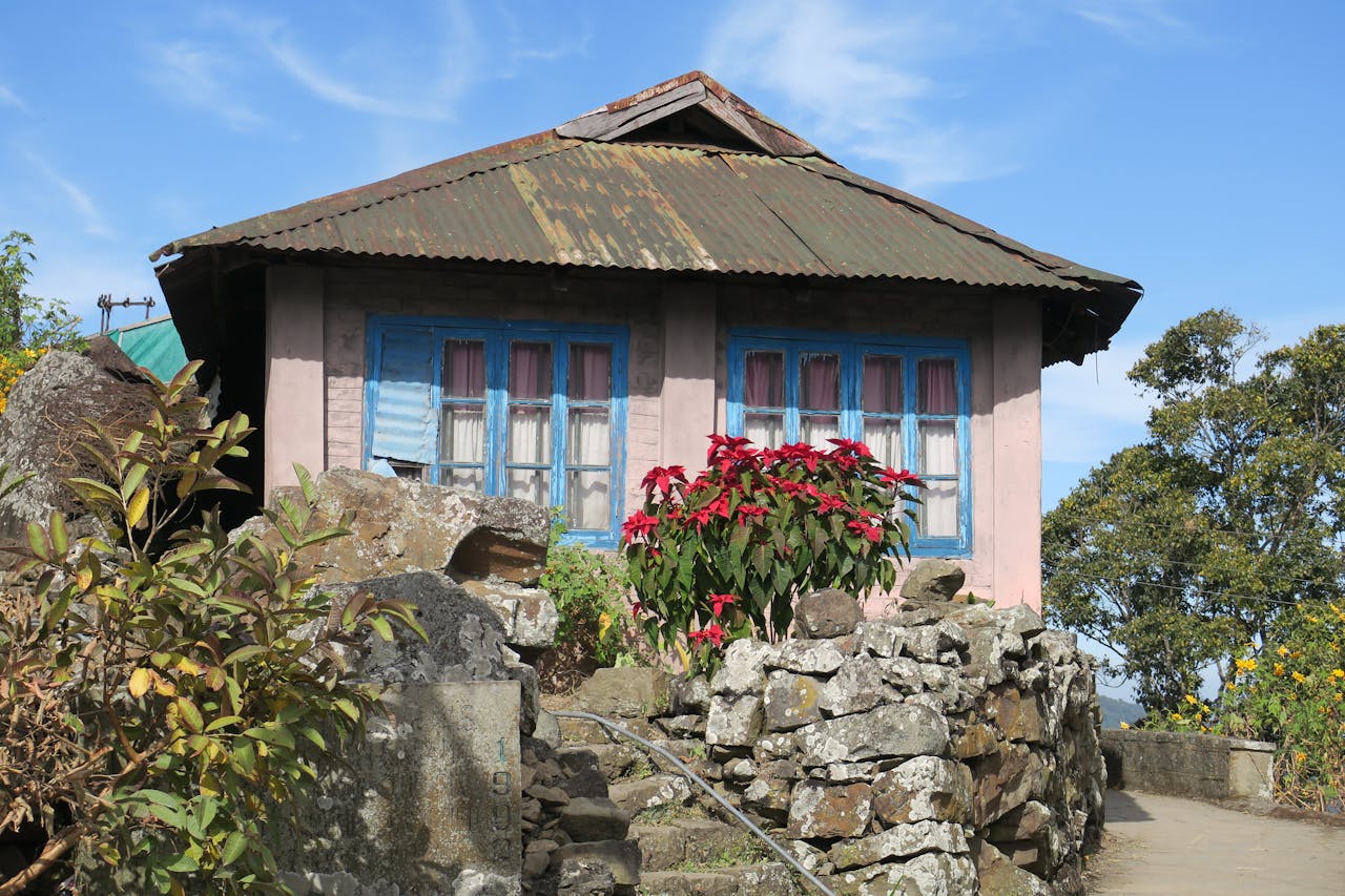 Charming rural house with a tin roof, stone wall, and vibrant flowers under a sunny sky.